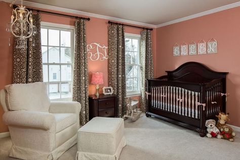 Warm, peachy-rose walls contrast with the dark wood crib and side table. An ivory armchair with a matching stool sits underneath a feminine, white chandelier. Brown and white patterned curtains make a funky statement. Dark Wood Crib, Nursery Dark Furniture, Transitional Nursery, Crib With Changing Table, Brown Crib, Wood Crib, Baby Nurseries, Wood Nursery, Trendy Baby Nursery