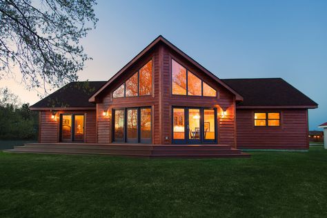 The rear of the home lit up at dusk.  You can see the large cathedral ceiling through the trapezoid windows and patio doors in the Great Room.  Master Bedroom patio door and kitchen window also lit up.  Cedar siding and other rustic accents. Chalet Windows, Trapezoid Windows, Lakehouse Plans, Pretty Windows, Zen Rooms, Woods House, Chalet Style Homes, Barbie Houses, Farm Cabin