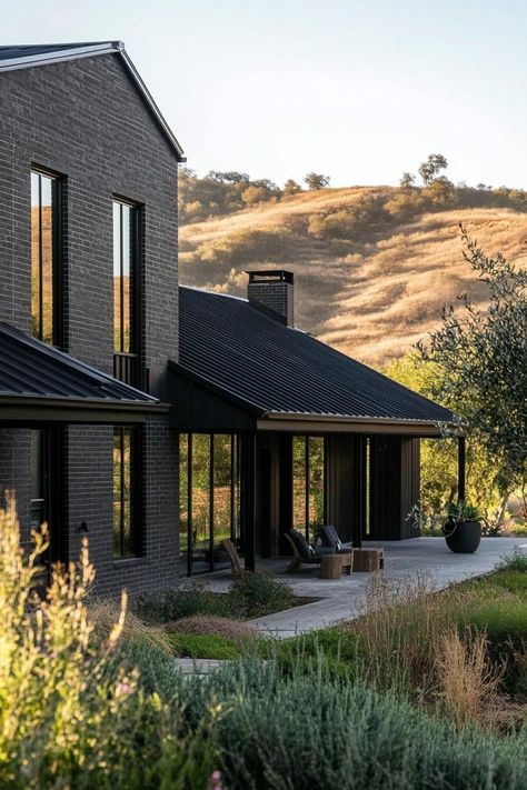 Modern barndominium home black brick facade and black metal roof stunning green hill landscape. Check out the coolest dark barndominium houses that combine rustic charm with modern sophistication, enhancing a living space with bold hues and sleek designs. Dark Barndominium, Barndominium Home, Barndominium Houses, Black Metal Roof, Hill Landscape, Black Window Frames, Modern Residential Architecture, Modern Barndominium, Black Houses