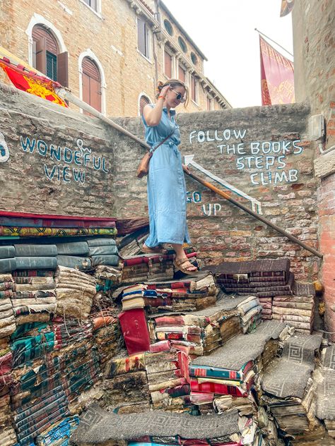 Woman in 20s wearing a light blue, ankle length button down dress walking down a staircase made of books at a famous bookstore in Venice, Italy. The books are water damaged. Her hair is in a claw clip and she’s wearing a brown leather cross body bag with Birkenstock sandals. Italian Bookstore, Italy Book Aesthetic, Outfit Inspo Italy, Venice Italy Aesthetic Vintage, Italy Bookstore, Book Staircase, Libraria Aqua Alta Venice, Liberia Acqua Alta, Venice Beach Graffiti