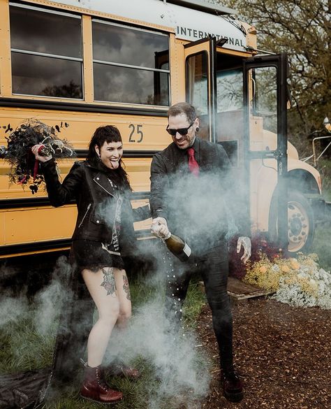 A tattooed couple standing outside a yellow American school bus. They are laughing and the bride is sticking her tongue out. The bride is wearing a Metallica t-shirt dress and holding a dark flower bouquet. The groom is wearing a black suit with red tie and popping a champagne bottle. The is a white smoke bomb smoking on the floor. Working With People, Alt Wedding, Rock N Roll Wedding, People Getting Married, Different Types Of People, Wedding Newspaper, Newspaper Design, Moody Wedding, Legal Documents