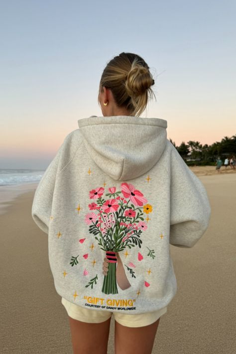 photo shows a blonde teenage girl standing on a sandy beach at sunset. she is facing away from the camera, looking at the ocean in the distance. she is wearing cream colored sweat shorts and a gray hoodie that has a big embroidered graphic of a bouquet of flowers on the back. "gift giving" and "courtesy of dandy worldwide" are embroidered on the back as well. the girl has her hair in a bun. the sky is orange and blue, like the sun is just about to set Dandy Flower Hoodie, Gift Giving Hoodie, Danny Worldwide Hoodie, Cute Aesthetic Hoodies, Embroidery Designs On Hoodies, Hoodies That Hoodie, Dandy Worldwide Hoodie, Things For Christmas List, How To Style A Hoodie