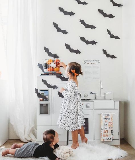 A child stands by a play kitchen, reaching towards festive decorations on a shelf, while another child sits on the floor nearby. The wall is adorned with bat silhouettes and a framed text. The scene is set in a bright room with white curtains. Bats Vampire, Kids Toy Kitchen, Vampire Bats, Toy Kitchen Set, Love At First Bite, Play Kitchens, Vampire Halloween, Vampire Bat, Kids Kitchen