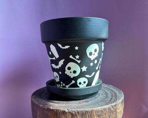 From the depths of darkness, a full moon rises and casts a haunting glow. Perfect for a haunted garden, this planter is made complete with skulls and bones, bats, coffins and stars.   Glow in the dark black or purple planter: * Terracotta Pot for indoor plants * Glow in the dark permanent vinyl * Comes with matching black tray * Surprise spooky gift included  *THE PLACEMENTS MIGHT BE A LITTLE DIFFERENT SINCE ALL DESIGNS ARE UNIQUE AND HANDMADE.* Care instructions: Wipe vinyl clean with a damp cl Halloween Flower Pots, Skeleton Planter, Halloween Pots, Haunted Garden, Stars Glow In The Dark, Pastel Goth Decor, Skulls And Bones, Skull Planter, Paint Inspo