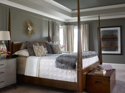 A tray ceiling painted with light and dark shades of gray and trimmed in white creates dimension over this stylish bedroom design. A wooden bed frame features tall pyramid posts, and the bed linens combine a traditional white cover with a faux fur accent throw and decorative pillows. A metallic sunburst draws light to the wall to finish the look. Paint With Wood Trim, Bedroom Wood Bed, Painted Tray Ceilings, Tray Ceiling Paint Ideas, Bedroom Tray Ceiling, Tray Ceiling Bedroom, Bedroom With Tray Ceiling, Tray Ceiling Ideas, Trey Ceilings