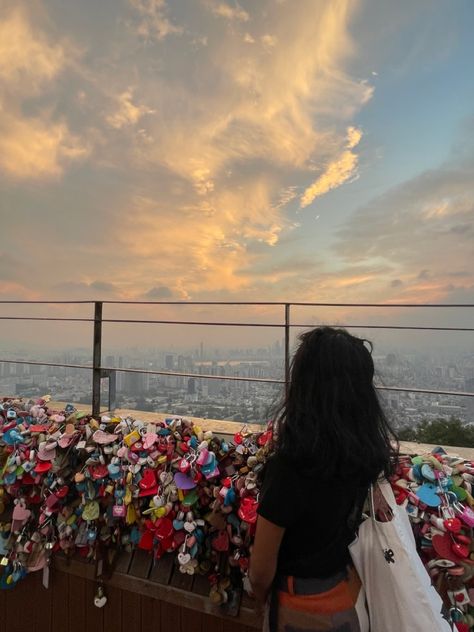 woman overlooking sunset, back facing towards the camera. She is wear a black shirt and patchwork pants. She is facing a railing covered in locks. South Korea Study Abroad, N Seoul Tower Aesthetic, Summer In Korea Aesthetic, Seoul In Summer, South Korea Seoul Aesthetic Outfit, Seoul Instagram Story, South Korea Trip Aesthetic, Study Abroad Japan, Study Abroad Aesthetic Korea