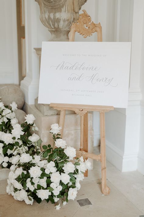 A landscape wedding welcome sign printed on a white background with grey calligraphy and text. The sign is displayed on an ornate wooden carved easel at the bottom of a small flight of stairs. An arrangement of white roses is displayed around the foot of the easel and a large stone urn stands behind the easel. Wedding Sign Easel, Wedding Signage Table, Wedding Table Signage, Sign Easel, Flowers And Pampas, Event Signs, Suzanne Neville, Event Signage, Wooden Easel