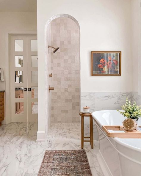 This cream and white bathroom features a hidden doorless walk-in shower behind a wall partition, a white tub, and marble flooring. A small table accessorized bath salts rest near the tub. A dark, traditional runner stretches along the floor below the soaking tub. Hidden Shower, Small Master Bath, Doorless Shower, Master Bath Shower, Window In Shower, White Tub, Master Shower, Master Bath Remodel, Upstairs Bathrooms