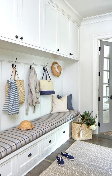 Gray bi-fold doors open to a cream colored rug placed in front of a long white bench accented with oil rubbed bronze cup pulls and a yellow and gray striped bench cushion topped with gray and cream pillows placed against a white shiplap trim beneath iron hooks fixed beneath white overhead cabinets. Farmhouse Mudroom, Mud Room Entry, Mudroom Entryway, Mudroom Decor, Mudroom Laundry Room, Mudroom Design, Mud Room Storage, Diy Entryway, Bench Designs