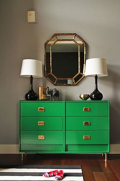 Gorgeous bedroom with gray walls framing a emerald green dresser with brass campaign style hardware and brass legs topped with a pair of glossy black table lamps, a gold vase and brass bowl below a gold faux bamboo mirror over hardwood floors layered with a black and white striped rug. Ikea Rast Hack, Gray Bedroom Walls, Campaign Dresser, Dresser Black, Green Dresser, Gorgeous Bedrooms, Mirror On The Wall, Bedroom Dresser, Trendy Bedroom