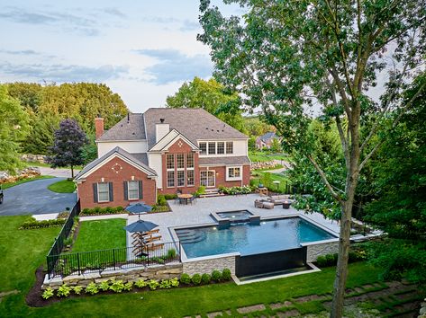 Isn't she lovely? Dominion features a negative edge, spa with an overflow, and dark grey pool tile transforming this traditional rectangular pool into a jaw-dropping work of art. With its dark, moody water and dramatic overflow cascading down a stone veneer retaining wall, the space feels sophisticated and opulent with a pool as beautiful as it is functional. Pool On Sloped Yard Retaining Walls, Inground Pool And Fire Pit Ideas, Pool With Wall On One Side, Pool Perpendicular To House, Red Brick House With Pool, Rectangle Pool With Retaining Wall, Sloped Yard Pool, Grey Pool Tile, Mobile Fire Pit