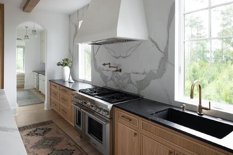 A white range hood is fixed against a honed marble slab backsplash between windows and over an aged brass swing arm pot filler and a stainless steel dual range. Drywall Range Hood, White Kitchen Drawers, Marble Slab Backsplash, Northern Preppy, Kitchen Range Hood Ideas, White Range Hood, Marble Top Kitchen Island, Black Marble Countertops, White Stove