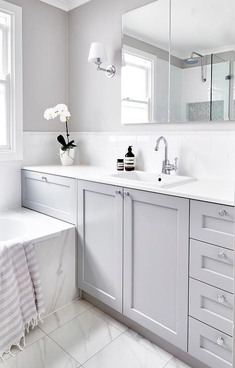 Beautiful gray and white bathroom is fitted with a gray washstand adorning polished nickel knobs and a white quartz countertop holding an overmount sink with a polished nickel gooseneck faucet in front of white subway backsplash tiles tiled with white pencil tiles fixed beneath a vanity mirror flanked by nickel sconces mounted on a gray wall. Grey Bathroom Paint, Grey Bathroom Cabinets, Makeover Kamar Mandi, Gray And White Bathroom, Grey Bathroom Tiles, Sconces Bathroom, Grey Bathroom Vanity, Bad Inspiration, Trendy Bathroom