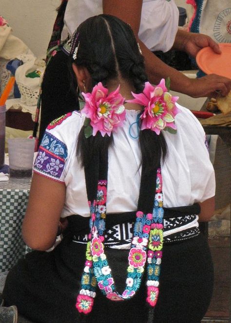 The cintas are black and embroidered in the design of cabbages. Also typical are the two large flowers entwined in her hair at the beginning of the braid. The designs vary both according to the taste of the person who did the embroidery, and the traditions of the village from which she came.  The ankle-length skirt is made of black wool, and is held up by a black and white embroidered belt. The overall effect is elegant. Traditional Mexican Hairstyles Braids, Mexican Braids With Ribbon, Mexican Hair Flowers, Braids With Ribbons In Them Mexican, Mexican Braids With Ribbon How To, Native American Braids, Native American Hair, Mexican Hairstyles, Pretty Braids