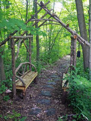 An arbor made of cedar wood with a seating area leads Wooded Yard Landscaping Ideas, Woods Path Ideas, Things To Do With Cedar Wood, Log Arbor, Woodland Paths Walkways, Forest Seating, Walk In The Woods Aesthetic, Wooded Pathway Ideas, Pathway In The Woods