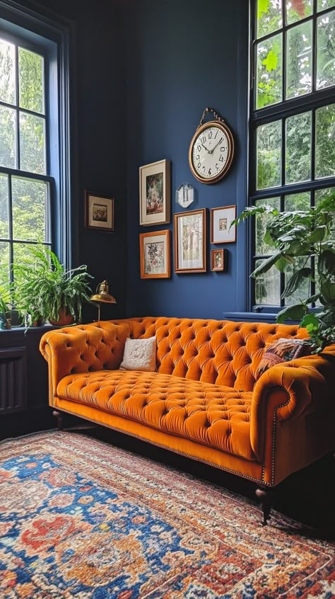 Bright orange velvet Chesterfield sofa in a cozy English living room with dark blue walls, vintage rug, and green plants. Orange Chesterfield Sofa, Interior Design Red Accents, Burnt Orange And Blue Living Room, Blue And Burnt Orange Living Room, Orange Sofa Decor Ideas, Blue Velvet Sofa Living Room Ideas, Dark Blue Sofa Living Room Ideas, California Cool Interior Design, Bold Living Room Ideas