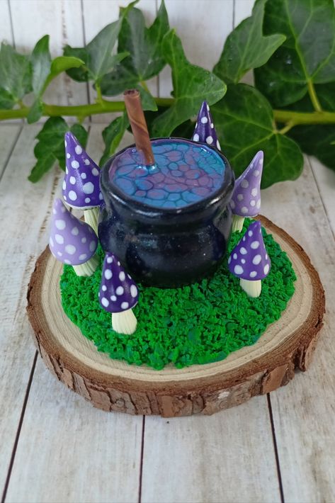 Image shows a miniature cauldron sculpted from polymer clay, with a shimemring blue and purple 'brew' inside. Surrounding the cauldron are little polymer clay toadstools in shades of purple, each with white spots. The cauldron and toadstools are mounted on a circular wooden slice with sculpted 'moss'. Black Cauldron, The Cauldron, The Black Cauldron, Wood Slices, Clay Projects, The Scene, Shades Of Purple, Polymer Clay, Shades