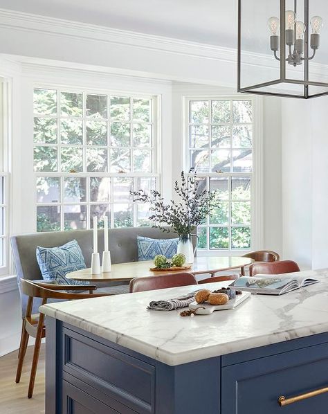 Bay window dining area boasts a gray settee placed at a gold oval dining table paired with brown wood dining chairs. Oval Breakfast Table, Window Breakfast Nook, Bay Window Breakfast Nook, White Dinning Room, Bay Window Dining, Settee Living Room, Saarinen Dining Table, Settee Dining, Cottage Dining Rooms