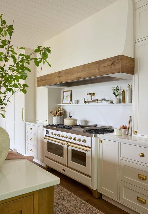 A brass swing arm pot filler is fixed to a marble slab backsplash between a white plaster hood with wood trim and a marble floating spice shelf, while white inset drawers with vintage brass knobs flank a white and gold French range. Hood With Wood Trim, French Range, White Cottage Kitchen, Oven Range Hood, Blue Laundry Rooms, Off White Kitchens, Wood Range Hood, Marble Shelf, Spice Shelf