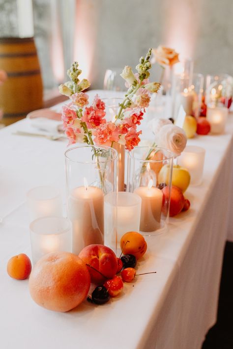 Using the colors of a peach as inspiration, this sweetheart table was filled with neutral colors so the brightness of the fruit and flowers stood out.  Taupe candles, colorful flowers and fruit across the front of the sweetheart table. Fruit Inspired Wedding Decor, Wedding Table With Fruit, Fruit As Wedding Decor, Fruit Table Decor Wedding, Wedding Centerpieces With Fruit, Fruit Wedding Table Decor, Fruit On Wedding Tables, Table Decor With Fruit, Fruit Wedding Centerpieces