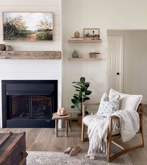 A brightly lit living room is designed with hardwood floors and a black fireplace. The chimney is wrapped in white shiplap with a rustic wood floating mantle mounted above. There are floating shelves next to fireplace that are decorated with a small painting, stacked books, and potted plants. A wood and white cushioned chair sits off to the side. Floating Mantle Shelf Living Room, Floating Shelf Next To Fireplace, Shelf Next To Fireplace, Floating Shelves Around Fireplace, Around Fireplace Ideas, Floating Shelves Next To Fireplace, Shelves Next To Fireplace, Shelves Around Fireplace, Floating Mantle