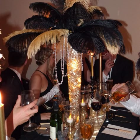 Large glass vase filled with gold scatter glass pieces and fairy lights, topped with black & gold ostrich feathers Feather Centre Piece Ideas, Gold Ostrich Feather Centerpiece, Black Ostrich Feather Centerpiece, Black And Gold Feather Centerpieces, Masquerade Ball Flower Arrangements, Roaring 20s Prom Decorations, Black N Gold Party Ideas, Black And Gold Prom Theme, Black And Gold Party Centerpieces