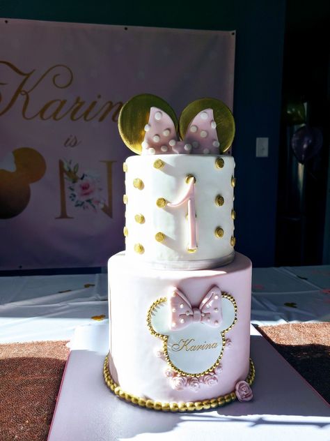 Two tier Minnie Mouse cake with gold Minnie ears on top and pink and white bow. Top tier shows child's age, and bottom tier shows child name surrounded by gold trimmed Minnie ears silhouette. Minnie Mouse Birthday Pink And Gold, Rose Gold Minnie Mouse Cake, Disney 1st Birthday Cake, Mini Mouse Cake 1st Birthday, Gold And Pink Minnie Mouse Cake, 1st Birthday Cake Minnie Mouse, Pink Minnie Cake, Pink Mini Mouse Cake, Minnie Cakes Birthday