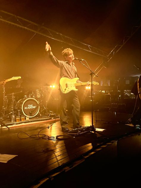 Craig Fitzgerald, the lead singer of the Academic is standing on stage in the golden spotlight with a guitar and one hand pointed in the air while standing in front of a microphone The Academic Band, Jess Aesthetic, Concert Wall, Academic Aesthetic, Silly Words, Hopelessly Devoted, Collage Images, 2024 Goals, Music Things