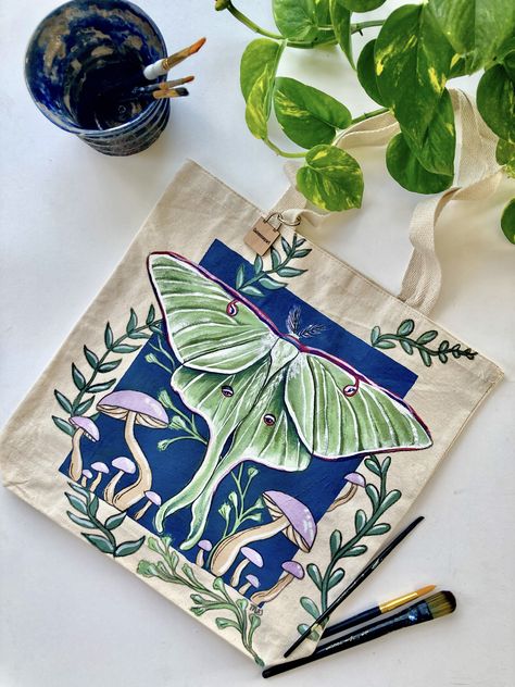 This is a green luna moth hand-painted tote bag with a navy blue background and purple mushrooms. Luna Moth Painting, Moth And Mushroom, Luna Moth Design, Diy Bag Painting, Diy Tote Bag Design, Painted Canvas Bags, Handpainted Tote Bags, Totes Ideas, Painted Clothes Diy