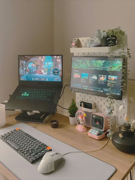 A compact work/gaming station featuring a laptop on a stand, a large monitor displaying a game, surrounded by playful decorations like miniature figures, a retro television, and various plants on a shelving unit beside the desk. The desk is adorned with a vintage lamp and other personal items, creating a cozy and inviting space. Computer Desk In Bedroom, Cozy Desk Setup, Positive Environment, Small Computer Desk, Cozy Desk, Clean Desk, Desk Setups, Small Gadgets, Desktop Setup