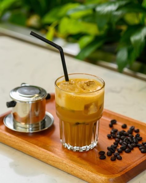 Alt text: "A glass of Cà Phê Sữa Đá (Vietnamese iced coffee with milk) served on a wooden tray alongside a traditional Vietnamese drip filter and scattered coffee beans, with lush greenery in the background." Vietnamese Iced Coffee, Egg Coffee, Coffee Shop Branding, Viet Food, Vietnamese Coffee, Blended Drinks, Instant Coffee, Coffee Grounds, Coffee Tea