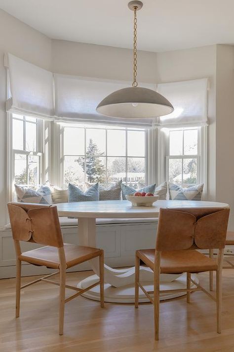 Brown leather dining chairs sit at a white oval dining table with a bay window dining bench lit by a gray lantern. Window Dining Bench, Bay Window Kitchen Ideas, Bay Window Dining Room, Brown Leather Dining Chairs, White Oval Dining Table, Bay Window Dining, Dining Table Design Ideas, Bay Window Benches, Kitchen Bay Window