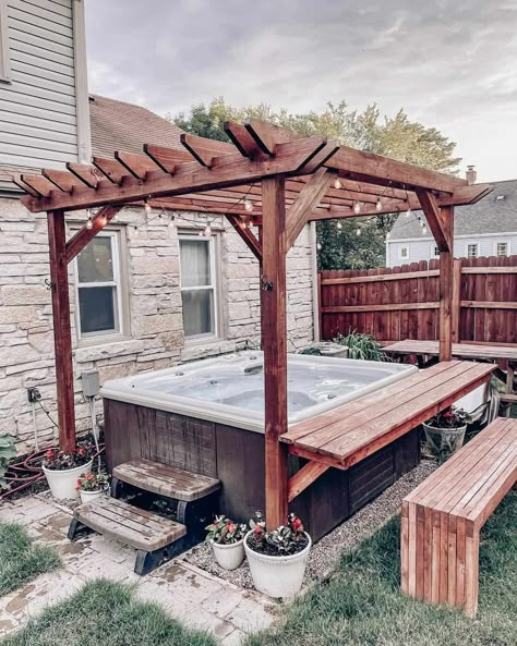 Take a soak in this brown hot tub covered by a stained wood canopy with black string lights. The canopy stands next to an adorable house made of beige stone. Make hanging out at the hot tub even easier with a wooden bar counter and wood bench. Hot Tub Ideas Backyard Pergola, Walkway To Hot Tub, Pergola Patio Hot Tub Ideas, Fenced In Hot Tub Area, Diy Hot Tub Table, Diy Hot Tub Pergola, Outdoor Patio Ideas With Hot Tub, Hot Tub Areas Outdoor, Hot Tub Porch Ideas