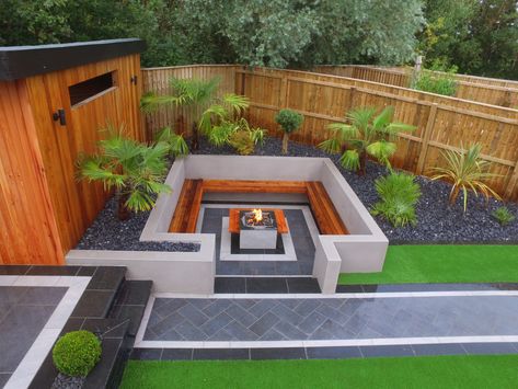 A stunning fire pit surrounded by bespoke built-in seating and contemporary grey garden paving made of porcelain. The fire pit sites beside the timber summer house Sunken Area In Garden, Angled Garden Design, Fire Pit Under Tree, Sunken Seating Area Garden, Sunken Fire Pit With Seating, Garden Fire Pit Ideas, Built In Garden Seating, Outdoor Fire Pit Ideas, Sunken Seating