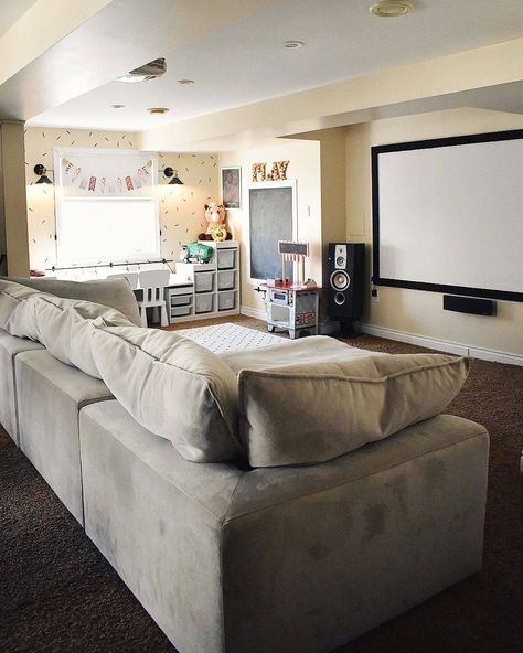 Family bonus room ideas are showcased in this space and include a gray sofa beside a small playroom. The playroom is equipped with white storage cubbies connected to a desk. The sofa sits in front of a large projector screen. Family Room Projector, Kids Media Room Ideas, Basement Playroom And Family Room, Teen Basement Hangout Ideas, Playroom Family Room Combo, Family Movie Night Ideas Living Rooms, Basement Playroom Ideas Family Room, Living Room And Playroom Combo, Family Bonus Room