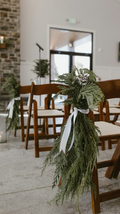 Spruce up your winter wedding with simple evergreen sprigs tied together with a white ribbon and hang on chairs in the aisle. Clean and elegant and smell so wonderful. Not too bulky for guests but adds a special winter touch to wedding day. Wedding Pine Decor, Pine Leaves Wedding Decor, Pine Bough Wedding Decor, Christmas Decor Chairs, Evergreen And Maroon Wedding, Wedding Arch Ideas Winter, Wedding In The Winter, Winter Wedding Welcome Table, December Wedding Arch