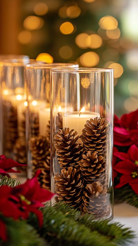 Glass candle jars with pine cones on a table adorned with red and green flowers, creating a cozy Christmas ambiance. Glass Cylinder Vases Christmas, Xmas Candle Decorations, Christmas Decor Glass Vase, Glass Bowl Christmas Centerpiece, Trendy Christmas Decor 2024, Christmas Votives Ideas, Easy Christmas Table Centerpieces Budget, Tall Clear Vase Filler Ideas, Christmas Ornament Centerpieces