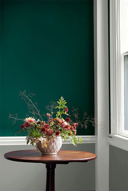 A hunter green wall with white trim and gray wainscoting frames a round wooden table on which a vase of flowers sits. Emerald Green Mudroom, Jewel Toned Green Paint, Jewel Tone Green Bedroom, Jewel Tone Entryway, Emerald Green Painted Walls, Benjamin Moore Accent Wall Colors, Jewel Green Paint Colors, Hunter Green Benjamin Moore, Hunter Green Dining Room