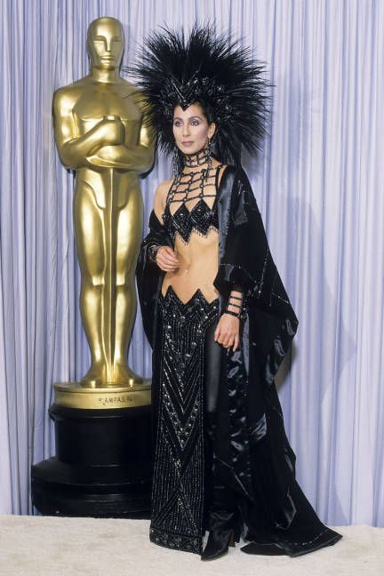 Cher at Oscars in 1986 (in midriff-bearing Bob Mackie getup featuring beaded loincloth, black cashmere cape, & mohowk-style headpiece made of hundreds of rooster feathers) Cher Black Outfit, Cher Bob Mackie, Cher 70s, Oscars Fashion, Cher Fashion, Cher Costume, Cher Outfits, Academy Awards Red Carpet, Cher Photos