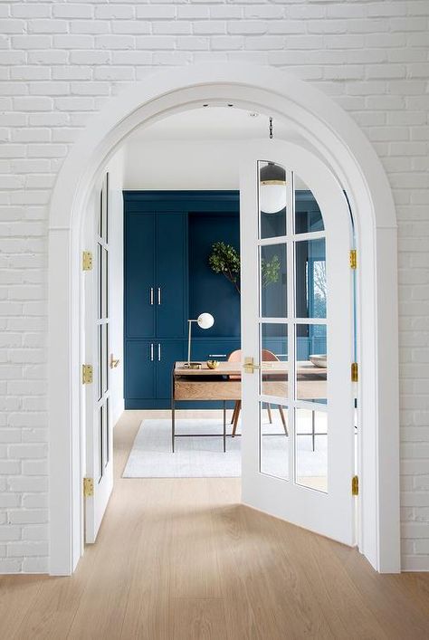 White brick frames arched French doors leading into a home office styled with a brown leather desk chair placed on a white rug at a wood and brass desk lit by a Hicks pendant. Home Office French Doors, Office Cabinet Design, Office French Doors, Green Home Offices, Arched French Doors, Den Library, Wood And Metal Desk, Home Office Shelves, Library Office