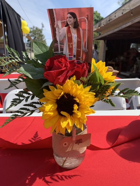 Mason jar . Mason jar with sunflowers , red carnations and red roses . Mason jar centerpiece . Fresh flowers . Burlap wrapped around the mason jar . USC colors red and yellow . Best part to add a picture of the celebrated graduate . Cute and super simple Graduation Party Centerpieces Mason Jars, Yellow Party Decorations, Red Roses Centerpieces, Floral Graduation Party, Red Centerpieces, Senior Graduation Party, Graduation Flowers, Mason Jar Centerpiece, Sunflower Party