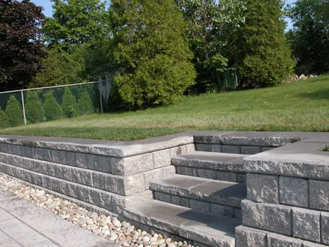 Steps and retaining wall accommodating to levels of patio Diy Retaining Wall With Steps, Retaining Wall Entrance, Versa Lok Retaining Wall, Parking Pad Retaining Wall, Steps In Retaining Wall, Patio With Retaining Wall Ideas, Sunken Patio Retaining Walls, Concrete Patio With Retaining Wall, Deck With Retaining Wall