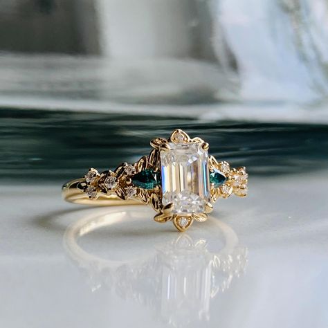 Emerald cut moissanites engagement ring in yellow gold. Fantasy ring. * 1ct white moissanites (5x7mm) * 14k gold Arwen setting * 12 accented moissanites * Free sizing * 7 days return policy This engagement ring features an emerald cut moissanite, sparkling and clean. It is set in our unique 14k Gold setting, 2 green and 10 clear moissanites on the sides. Immerse yourself in the whimsical beauty of the Arwen fantasy-inspired gold engagement ring! Let this extraordinary piece be a symbol of your love story, capturing the magic and wonder that brought you together. It is a ring that will forever hold a special place in your heart and become a cherished heirloom for generations to come. This ring would make a beautiful unique engagement ring or promise ring as well as anniversary gift. It come Emerald Engagement Ring Unique, Classy Engagement Ring Vintage, Green And Gold Engagement Ring, Fantasy Rings Engagement, One Of A Kind Engagement Rings, Fairy Rings Jewelry, Elven Engagement Ring, Engagement Rings Fairy, Whimsical Rings