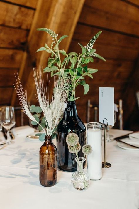 a boho wedding centerpiece of apothecary bottles, a candle, wheat and greenery for a simple look - Weddingomania Rustic Mountain Wedding Centerpieces, Greenery Vase Centerpiece, Wedding Centerpieces Bottles, Boho Mountain Wedding Decor, Pottery Centerpieces Wedding, Colorado Wedding Decor, Mountain Centerpieces Wedding, Mountain Cabin Wedding, Boho Ranch Wedding