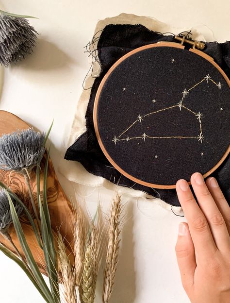 A hand holding a round embroidery hoop on a white surface. The hoop has navy fabric in it with a Leo constellation embroidered in silver fabric. There are stars embroidered around it. Beside the hoop is some wheat and blue flowers. Regulus Constellation, Leo Embroidery, Embroidered Constellations, Constellation Wall Art, Constellation Wall, Leo Constellation, Embroidered Hoop, Leo Birthday, Embroidery Sweater