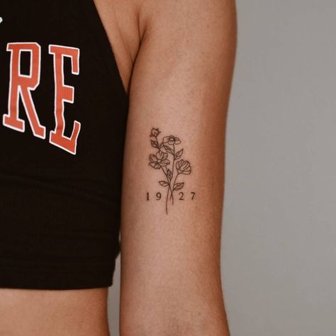 Flower bouquet and "1927" tattooed on the bicep. Fineline Bicep Tattoo, Flower Tattoo With Birth Date, Date Tattoo With Flower, Flower Tattoo With Numbers, Flower Tattoo Bicep Women, Birth Year Tattoo With Flower, Year Tattoo With Flowers, Inner Upper Bicep Tattoo Women, Women Biceps Tattoo