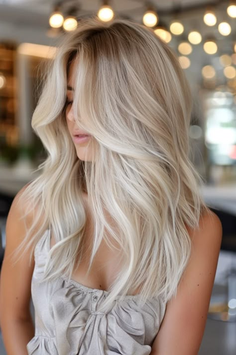 Woman with long, wavy blonde hair standing in a brightly lit room with decorative lights in the background. Level 7 Lived In Blonde, Champagne Blonde Hair Balayage Dark Roots, Milky Blonde Balayage, Cold Blonde Hair Balayage, Summer Blonde Hair Balayage, Blonde Balayage On Brunette Hair, Old Money Blonde Dark Roots, Bright Blonde Hair With Shadow Root, Highlights With Dark Roots