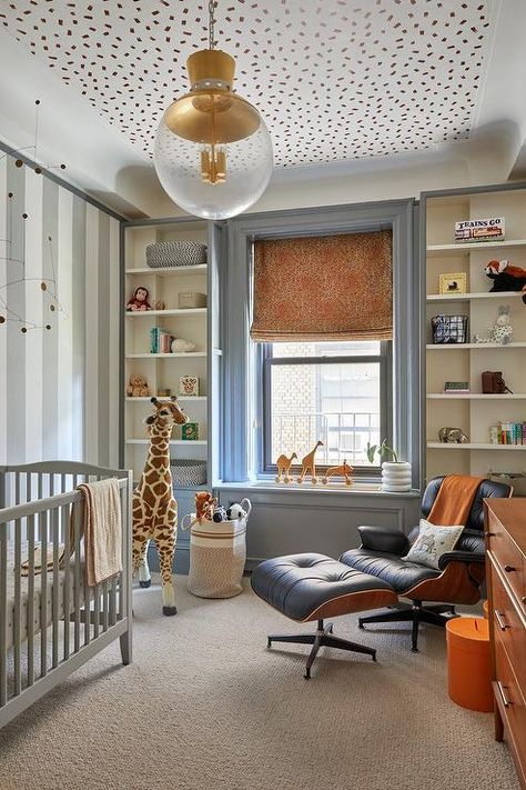 Boy's nursery features an Eames management chair and ottoman, a gray crib, a tall stuffed giraffe and built in shelves that surround a window under gold confetti ceiling wallpaper. Kids Wallpaper On Ceiling, Wallpaper Ceiling Boys Room, Wallpaper For Built Ins, Nursery Wallpaper On Ceiling, Post Modern Nursery, Built In Crib, Modern Eclectic Nursery, Grey Carpet Nursery, Nursery Ceiling Wallpaper