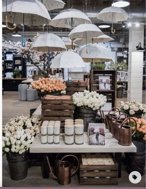 With the stunning color palette of white and orange, as well as the umbrella above the focus display, it's easy to imagine a stunning DIY fall retail display idea for this store. As a focal point, this table draws your attention to the area. The plants, candles, magazine, and water container make for a colorful and eye-catching display. This is probably the first thing I'd want to look at when I walk into the store. Boutique Window Displays, Spring Window Display, Magnolia Homes Paint, Gift Shop Displays, Painted Branches, Window Display Retail, Store Window Display, Focus Point, Store Window Displays