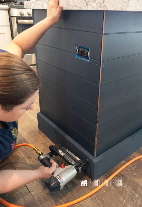 Woman holding a brad nailer secures the navy blue painted base molding to the bottom of the kitchen island. Kitchen Island With Shiplap, Island With Shiplap, Kitchen Island Remodel Ideas, Shiplap Bar, Kitchen Island Trim, Kitchen Island Base, Diy Kitchen Ideas, Shiplap Trim, Kitchen Island Makeover